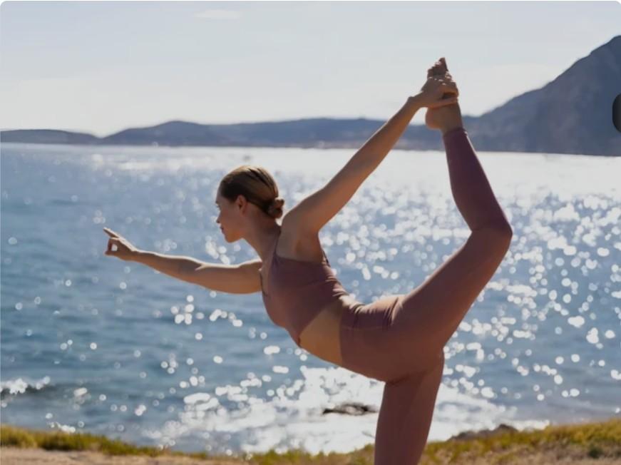 Woman practicing the King Dancer yoga pose by the ocean while wearing a mauve Ixora sports bra and matching leggings set.